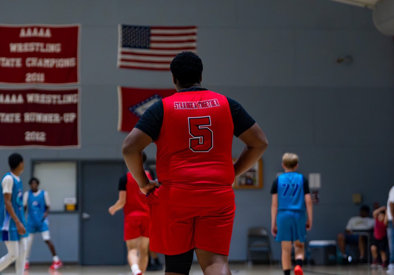 Basketball player in red jersey on indoor court, with American flag in background.